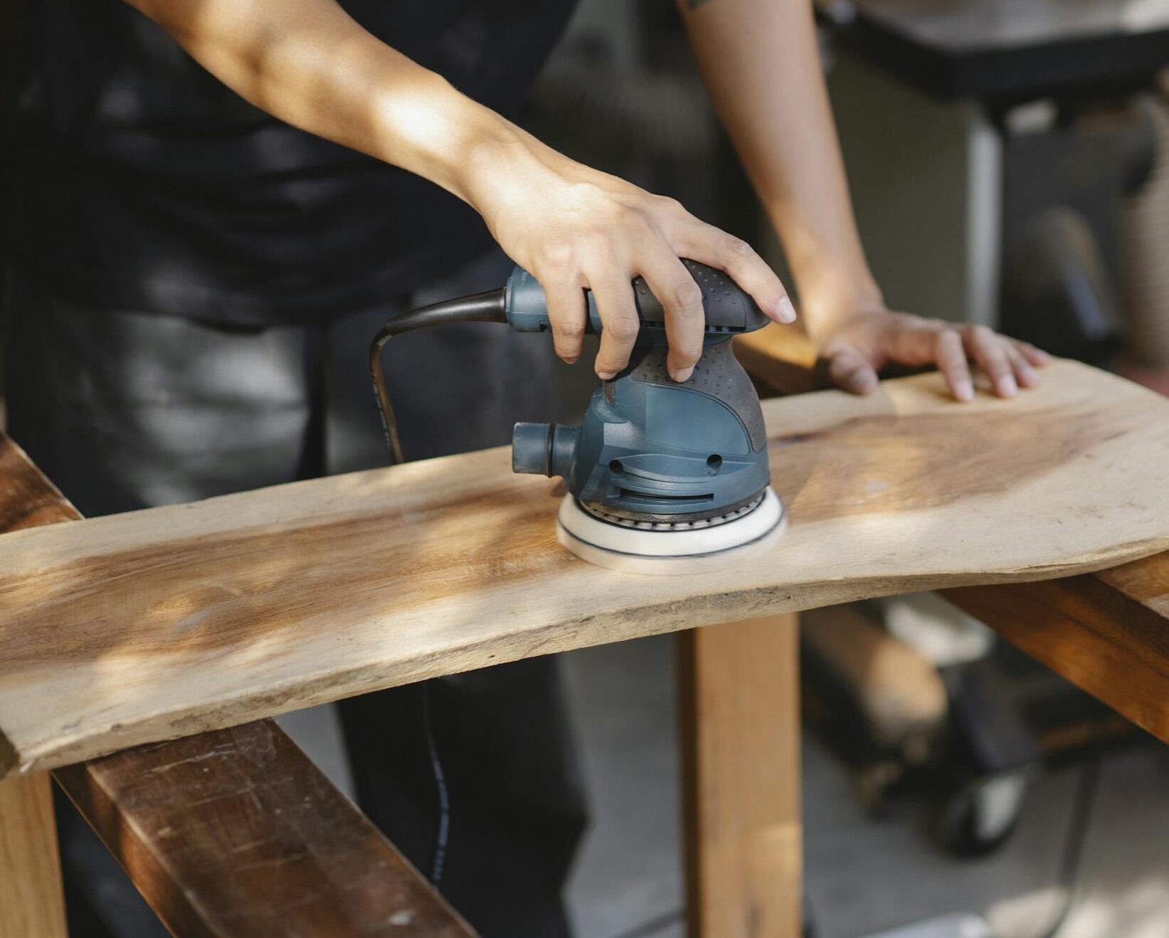 A craftsman uses a power sander on a wooden plank in a well-lit workshop.